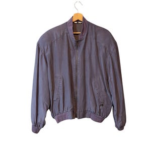 Könnte beinhalten: Vintage, staubig-lila Bomberjacke mit durchgehendem Reißverschluss. Die Jacke hat einen gerippten Kragen, Bündchen und Saum sowie zwei Vordertaschen. Sie hängt an einem Holzbügel vor weißem Hintergrund.