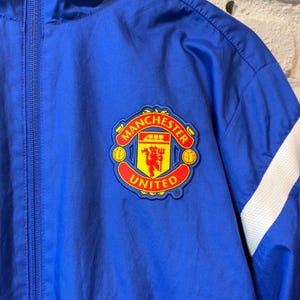 Puede incluir: Chaqueta azul con cremallera y parche del logo del Manchester United. La chaqueta tiene detalles blancos en las mangas y una cremallera completa. El logo presenta un diablo rojo y las palabras "Manchester United".
