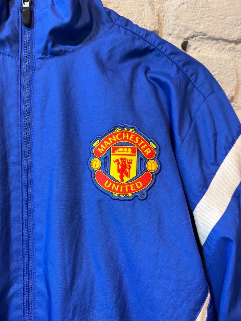 Puede incluir: Chaqueta azul con cremallera y parche del logo del Manchester United. La chaqueta tiene detalles blancos en las mangas y una cremallera completa. El logo presenta un diablo rojo y las palabras "Manchester United".