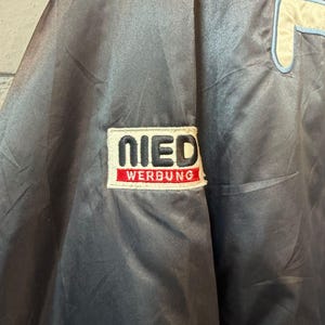 Op de afbeelding: Donkerblauwe satijnen jas met een groot cr&egrave;mekleurig en lichtblauw logo aan de rechterkant en een patch op de linkermouw. De patch leest "NIED WERBUNG" in witte letters op een rode achtergrond. De jas heeft een glanzende uitstraling.