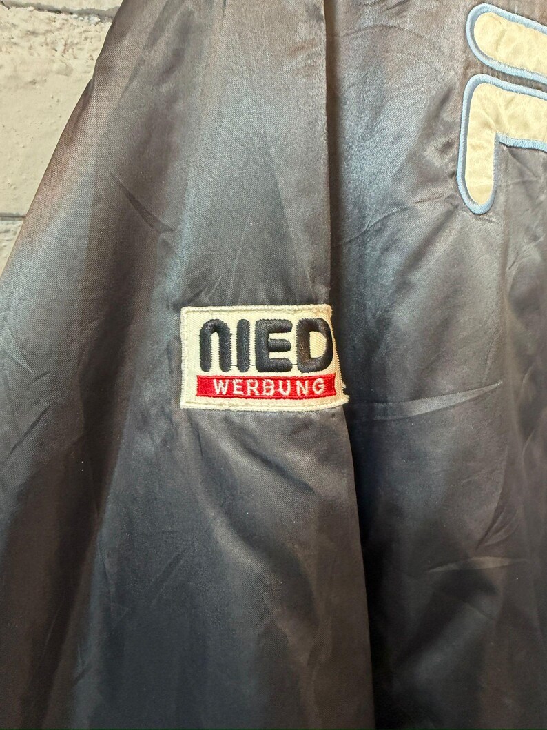 Op de afbeelding: Donkerblauwe satijnen jas met een groot cr&egrave;mekleurig en lichtblauw logo aan de rechterkant en een patch op de linkermouw. De patch leest "NIED WERBUNG" in witte letters op een rode achtergrond. De jas heeft een glanzende uitstraling.