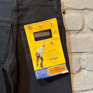 K&ouml;nnte beinhalten: Dunkelgraue Wrangler-Jeans aus Denim, die an einem Holzb&uuml;gel h&auml;ngen. Ein gelbes Etikett mit dem Wrangler-Logo und Text ist an der Ges&auml;&szlig;tasche befestigt. Das Etikett lautet "14 OZ. HEAVY DENIM" und "PRE-SHRUNK".