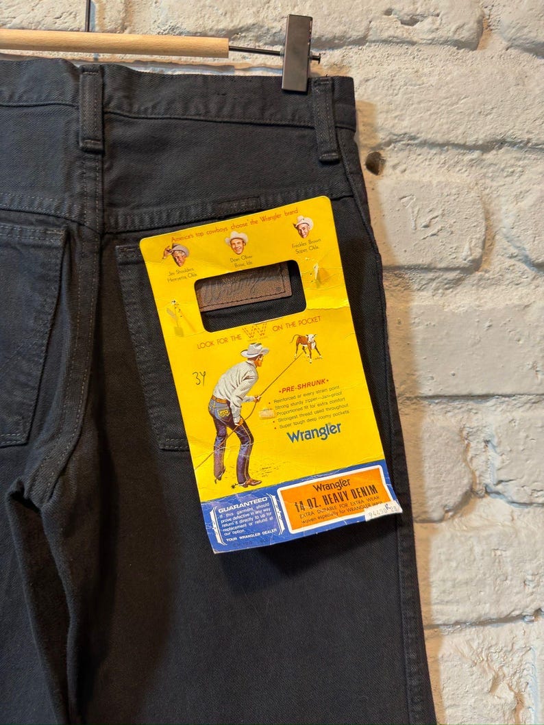 K&ouml;nnte beinhalten: Dunkelgraue Wrangler-Jeans aus Denim, die an einem Holzb&uuml;gel h&auml;ngen. Ein gelbes Etikett mit dem Wrangler-Logo und Text ist an der Ges&auml;&szlig;tasche befestigt. Das Etikett lautet "14 OZ. HEAVY DENIM" und "PRE-SHRUNK".