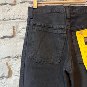 K&ouml;nnte beinhalten: Nahaufnahme einer schwarzen Jeans, die an einem Holzb&uuml;gel h&auml;ngt. Die Jeans hat ein klassisches F&uuml;nf-Taschen-Design mit einer Ges&auml;&szlig;tasche mit einem aufgestickten Design. Ein gelbes Etikett ist an der Jeans befestigt.