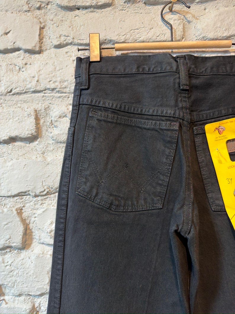 K&ouml;nnte beinhalten: Nahaufnahme einer schwarzen Jeans, die an einem Holzb&uuml;gel h&auml;ngt. Die Jeans hat ein klassisches F&uuml;nf-Taschen-Design mit einer Ges&auml;&szlig;tasche mit einem aufgestickten Design. Ein gelbes Etikett ist an der Jeans befestigt.