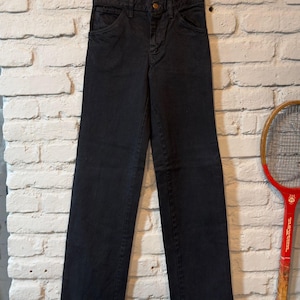 K&ouml;nnte beinhalten: Eine schwarze Jeans aus Denim, die an einem Holzb&uuml;gel h&auml;ngt. Die Jeans hat ein klassisches F&uuml;nf-Taschen-Design und einen Knopfverschluss. Die Jeans ist gerade geschnitten und scheint Vintage zu sein, vor einer wei&szlig;en Backsteinmauer.