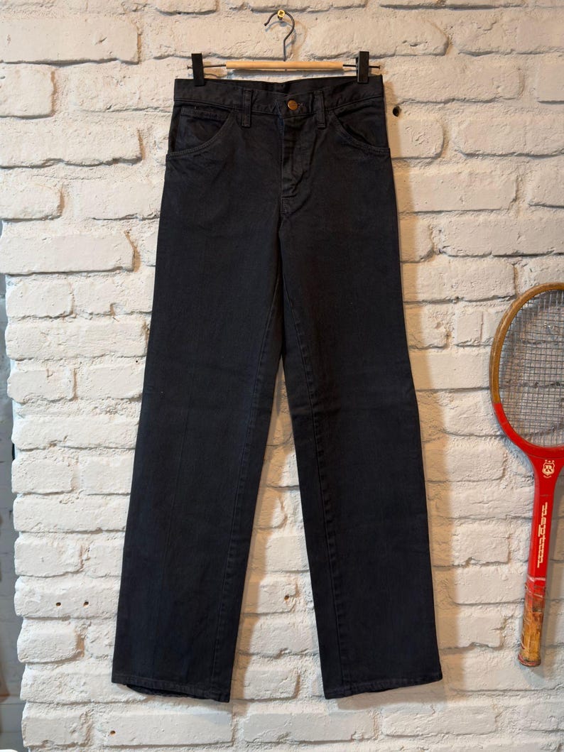 K&ouml;nnte beinhalten: Eine schwarze Jeans aus Denim, die an einem Holzb&uuml;gel h&auml;ngt. Die Jeans hat ein klassisches F&uuml;nf-Taschen-Design und einen Knopfverschluss. Die Jeans ist gerade geschnitten und scheint Vintage zu sein, vor einer wei&szlig;en Backsteinmauer.