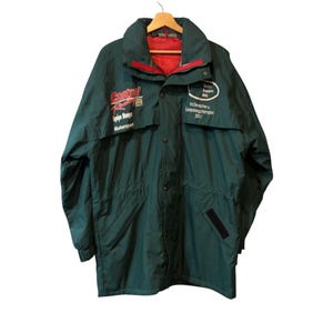 May include: A dark green jacket with a red lining and collar. The jacket has a high collar and a zip closure. The front of the jacket features the Castrol Racing logo and other text. The jacket is hanging on a wooden hanger.