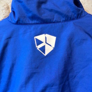 Puede incluir: Primer plano del cuello de una chaqueta azul brillante con un logotipo geom&eacute;trico blanco en la espalda. La chaqueta est&aacute; colgada de una percha de madera sobre una pared de ladrillos.