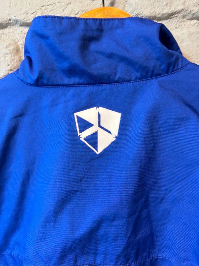 Puede incluir: Primer plano del cuello de una chaqueta azul brillante con un logotipo geom&eacute;trico blanco en la espalda. La chaqueta est&aacute; colgada de una percha de madera sobre una pared de ladrillos.