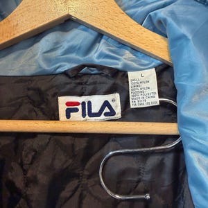 Op de afbeelding: Lichtblauwe FILA-jas aan een houten kleerhanger. De jas heeft een zwarte gewatteerde voering en een wit label met het FILA-logo en wasvoorschriften. De buitenkant, voering en vulling zijn gemaakt van nylon en polyester.