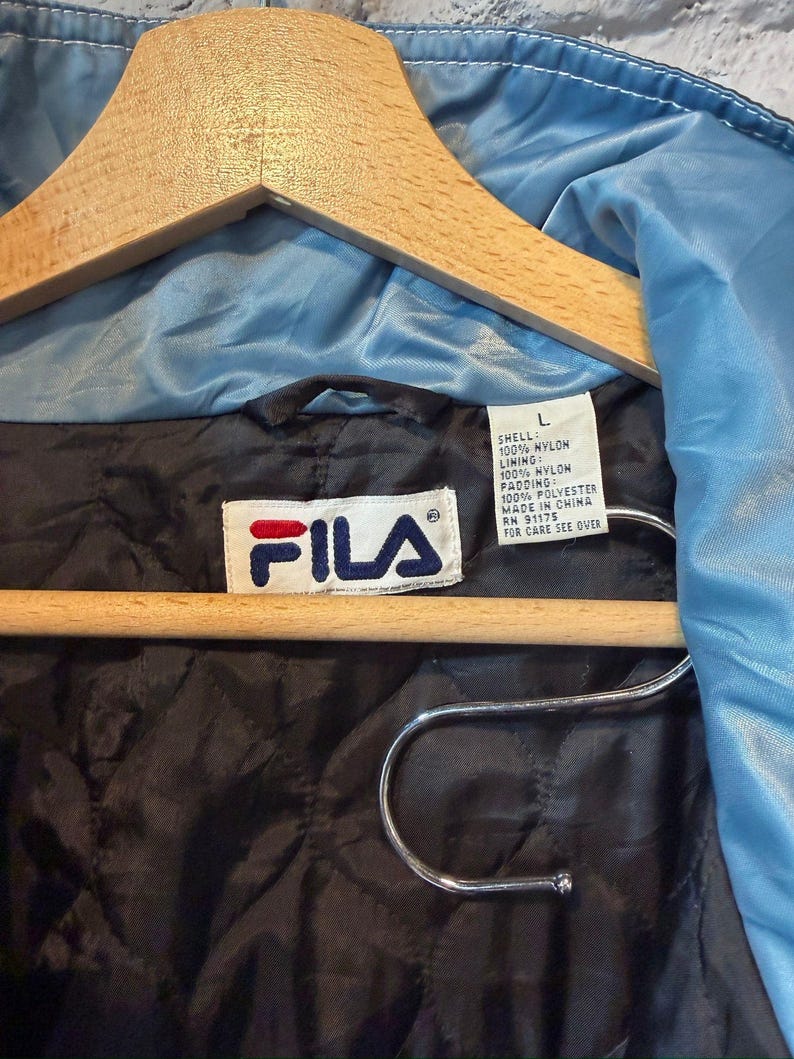 Op de afbeelding: Lichtblauwe FILA-jas aan een houten kleerhanger. De jas heeft een zwarte gewatteerde voering en een wit label met het FILA-logo en wasvoorschriften. De buitenkant, voering en vulling zijn gemaakt van nylon en polyester.
