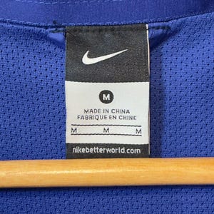 Puede incluir: Primer plano de una etiqueta de chaqueta deportiva azul con el logotipo de Nike y la letra "M". La etiqueta tambi&eacute;n incluye el texto "MADE IN CHINA FABRIQUE EN CHINE" y "nikebetterworld.com". La chaqueta est&aacute; colgada en una percha de madera.