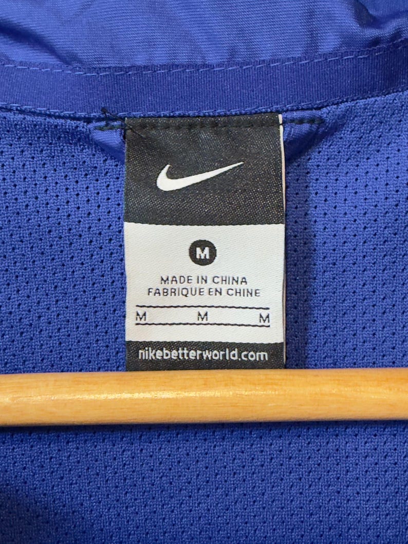 Puede incluir: Primer plano de una etiqueta de chaqueta deportiva azul con el logotipo de Nike y la letra "M". La etiqueta tambi&eacute;n incluye el texto "MADE IN CHINA FABRIQUE EN CHINE" y "nikebetterworld.com". La chaqueta est&aacute; colgada en una percha de madera.