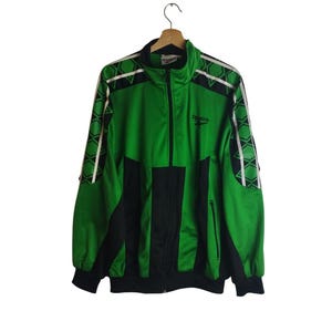 Pode incluir: Casaco de treino vintage verde e preto com fecho de correr completo. O casaco apresenta detalhes brancos nas mangas e ombros, e o logótipo da Reebok é visível no peito. Pendurado num cabide de madeira.
