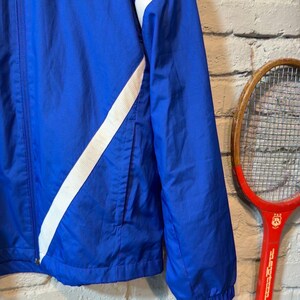 Puede incluir: Chaqueta azul con rayas blancas diagonales y el logo del Manchester United. La chaqueta tiene cierre de cremallera, bolsillo delantero y pu&ntilde;os el&aacute;sticos. Un logo del Manchester United es visible. Una raqueta de tenis roja vintage est&aacute; en el fondo.