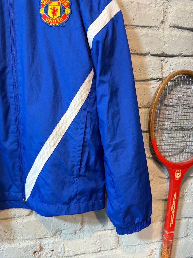 Puede incluir: Chaqueta azul con rayas blancas diagonales y el logo del Manchester United. La chaqueta tiene cierre de cremallera, bolsillo delantero y pu&ntilde;os el&aacute;sticos. Un logo del Manchester United es visible. Una raqueta de tenis roja vintage est&aacute; en el fondo.