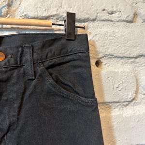 K&ouml;nnte beinhalten: Nahaufnahme einer schwarzen Jeans aus Denim, die an einem Holzb&uuml;gel h&auml;ngt. Die Jeans hat ein klassisches F&uuml;nf-Taschen-Design mit Knopfverschluss und einer kleinen Tasche auf der Vorderseite. Der Stoff scheint ein dunkles Denim zu sein.