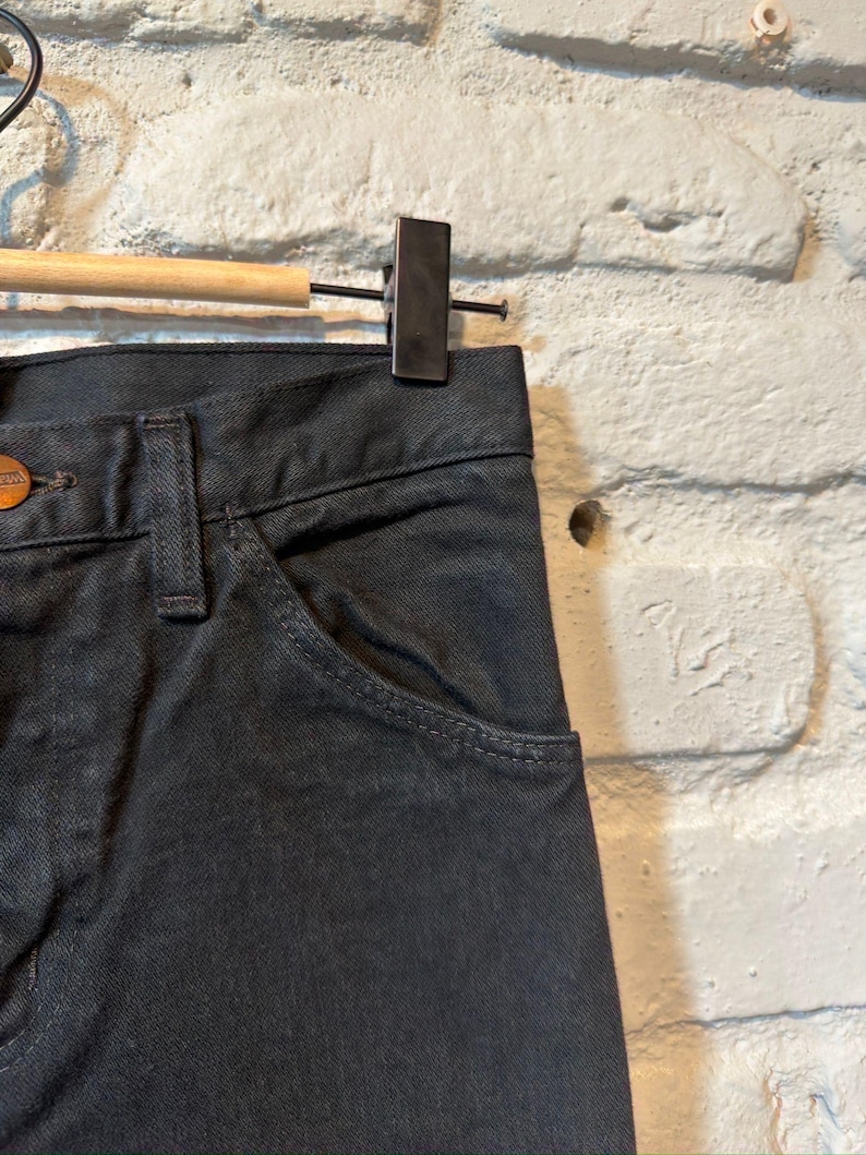 K&ouml;nnte beinhalten: Nahaufnahme einer schwarzen Jeans aus Denim, die an einem Holzb&uuml;gel h&auml;ngt. Die Jeans hat ein klassisches F&uuml;nf-Taschen-Design mit Knopfverschluss und einer kleinen Tasche auf der Vorderseite. Der Stoff scheint ein dunkles Denim zu sein.