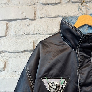 Op de afbeelding: Zwarte satijnen jas met een lichtblauwe voering, witte stiksels en een knoopsluiting. De jas heeft een patch met een berenafbeelding en de tekst "Die B&auml;ren Neuwied". De jas hangt aan een houten kleerhanger.