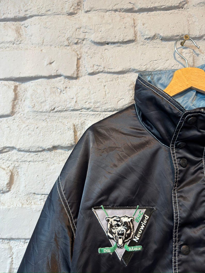 Op de afbeelding: Zwarte satijnen jas met een lichtblauwe voering, witte stiksels en een knoopsluiting. De jas heeft een patch met een berenafbeelding en de tekst "Die B&auml;ren Neuwied". De jas hangt aan een houten kleerhanger.