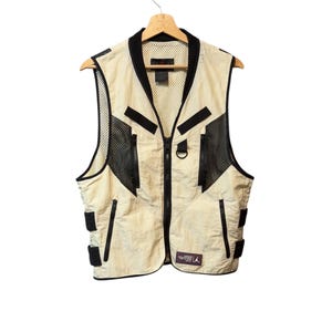 Op de afbeelding: Een beige vest met zwarte accenten, een ritssluiting over de hele lengte en meerdere zakken. Het vest heeft mesh details en een kleine logopatch. Het hangt aan een houten hanger tegen een witte achtergrond.