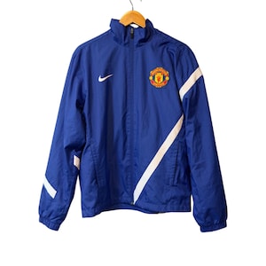 Puede incluir: Chaqueta azul Nike con rayas diagonales blancas en las mangas. Tiene cremallera completa, un peque&ntilde;o logotipo de Nike y el escudo del Manchester United en el pecho.