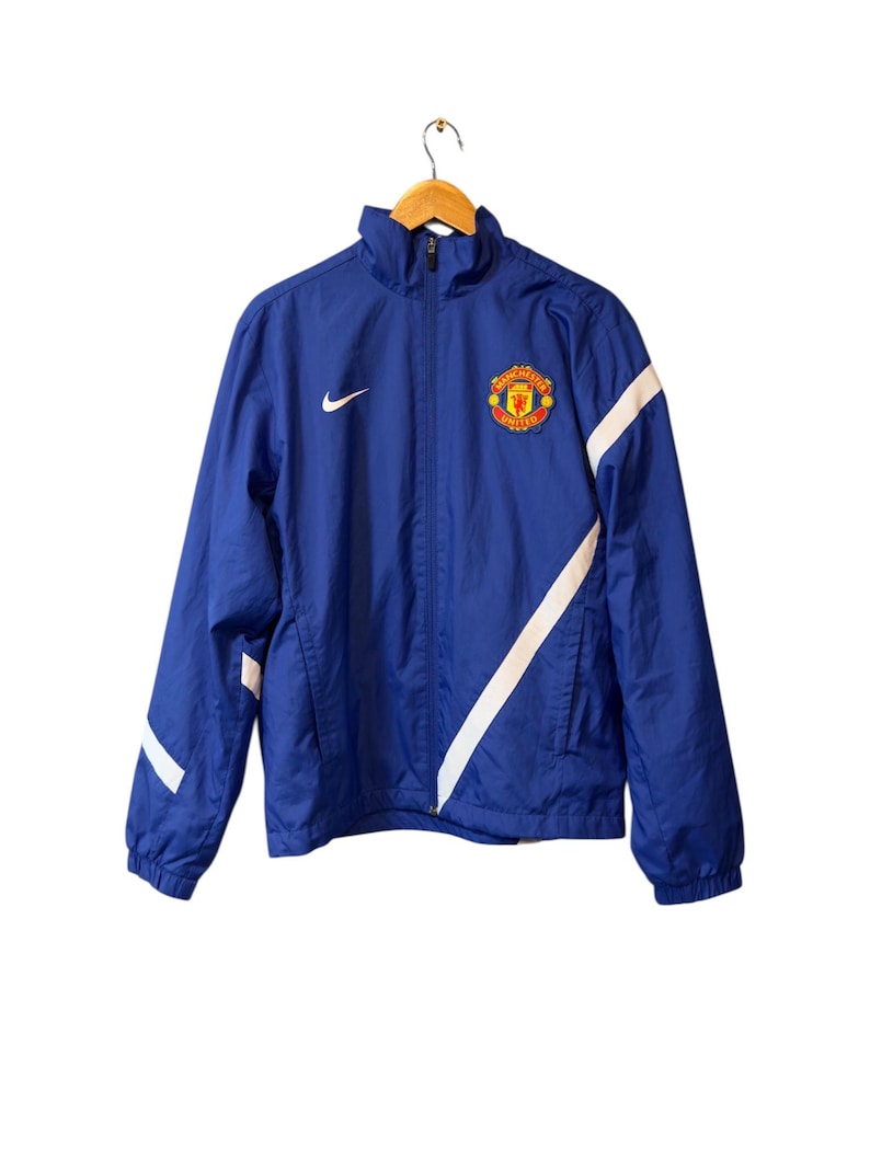 Puede incluir: Chaqueta azul Nike con rayas diagonales blancas en las mangas. Tiene cremallera completa, un peque&ntilde;o logotipo de Nike y el escudo del Manchester United en el pecho.