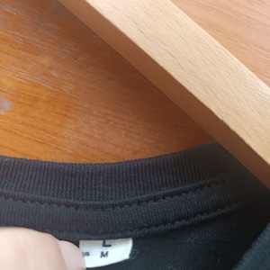 May include: Close-up of a black t-shirt collar with a white tag. The tag displays the sizes "L" and "M". The shirt is hanging on a wooden hanger.