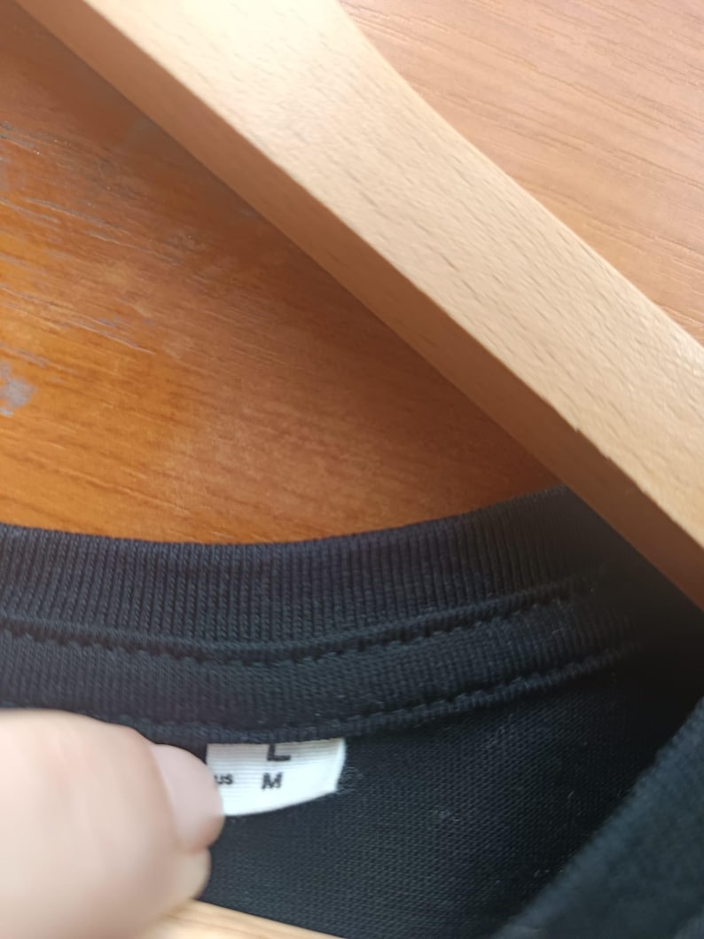 May include: Close-up of a black t-shirt collar with a white tag. The tag displays the sizes "L" and "M". The shirt is hanging on a wooden hanger.