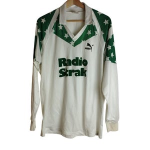 Puede incluir: Camiseta blanca de manga larga con estrellas verdes en los hombros y el cuello. La camiseta presenta el texto "Radio Strak" en letras verdes y un logotipo de Puma. La camiseta está colgada de una percha de madera.