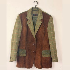 Può includere: Blazer in camoscio marrone con dettagli a quadri verdi sulle maniche, sul colletto e sulle tasche. Il blazer ha una chiusura a due bottoni. Il marchio "ADMONT" è visibile.