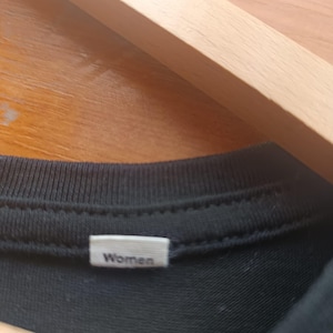 May include: Close-up of a black t-shirt collar with a white tag that reads "Women". The shirt is hanging on a wooden hanger.