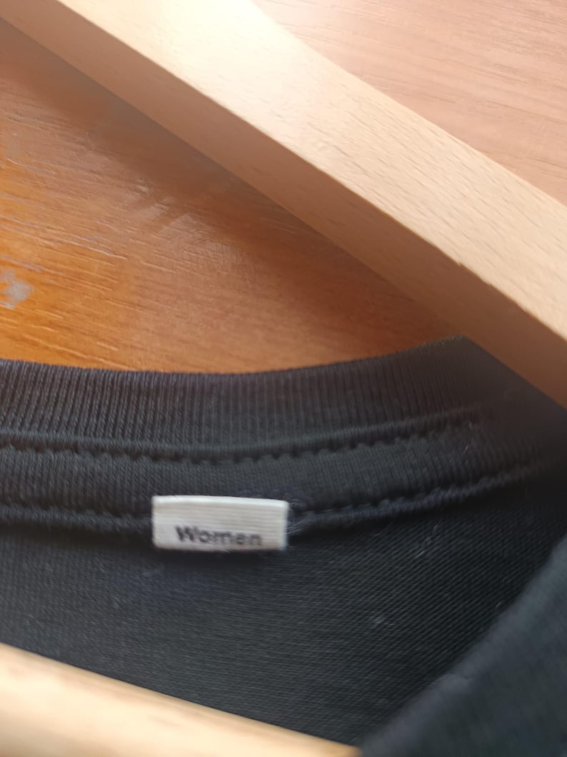 May include: Close-up of a black t-shirt collar with a white tag that reads "Women". The shirt is hanging on a wooden hanger.