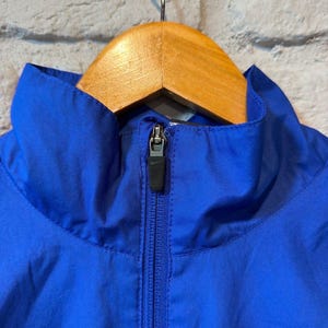 Puede incluir: Primer plano de una chaqueta azul vibrante con cuello alto y cremallera completa. La chaqueta est&aacute; colgada de una percha de madera. La cremallera es negra y la chaqueta parece estar hecha de un material ligero.