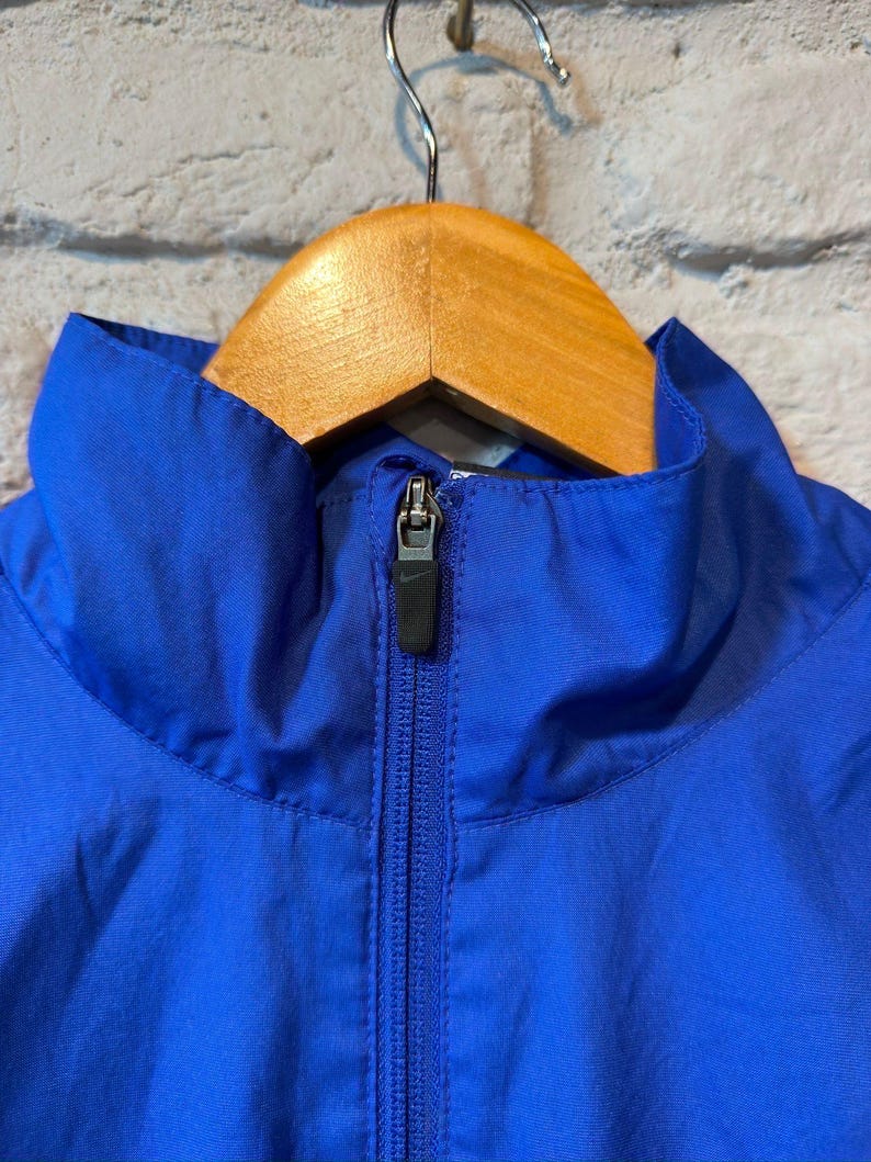 Puede incluir: Primer plano de una chaqueta azul vibrante con cuello alto y cremallera completa. La chaqueta est&aacute; colgada de una percha de madera. La cremallera es negra y la chaqueta parece estar hecha de un material ligero.