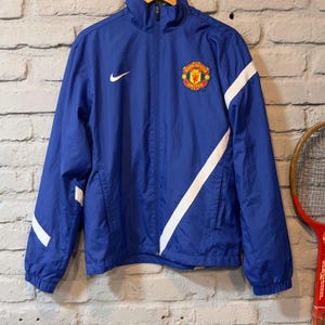 Puede incluir: Chaqueta azul con cremallera y rayas diagonales blancas, con el logo del Manchester United. El logo de Nike es visible en el pecho. La chaqueta cuelga de una percha de madera contra una pared de ladrillos.