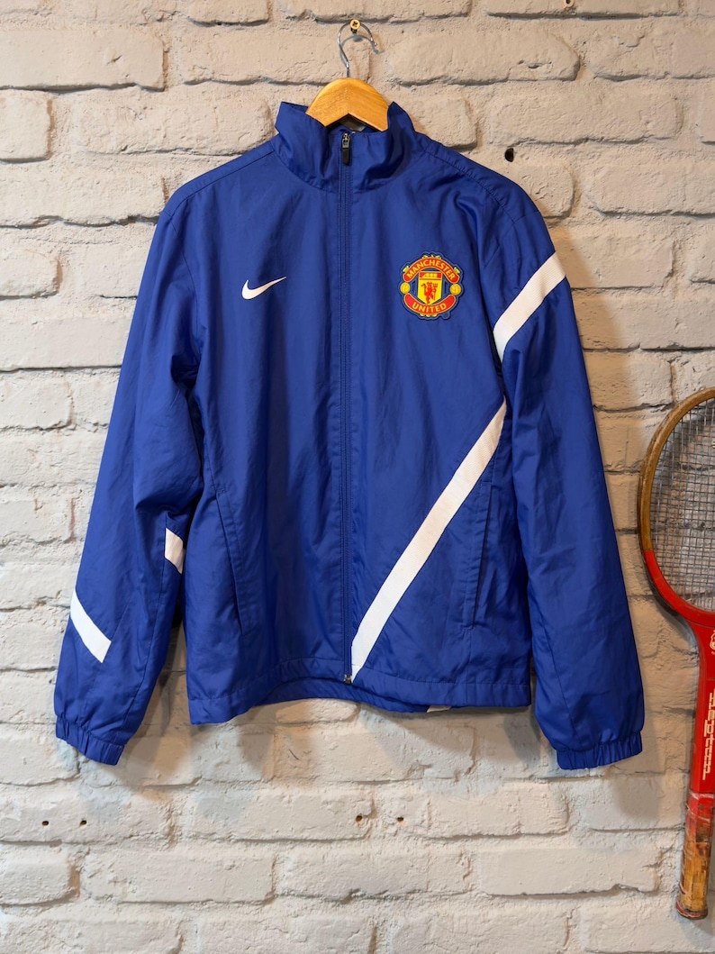 Puede incluir: Chaqueta azul con cremallera y rayas diagonales blancas, con el logo del Manchester United. El logo de Nike es visible en el pecho. La chaqueta cuelga de una percha de madera contra una pared de ladrillos.