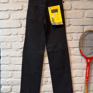 K&ouml;nnte beinhalten: Schwarze Jeans aus Denim, die an einem Metallb&uuml;gel vor einer wei&szlig;en Backsteinmauer h&auml;ngen. Die Jeans haben einen geraden Beinabschluss und ein gelbes Etikett mit einer Grafik einer Person. Ein Vintage-Tennisschl&auml;ger ist rechts zu sehen.