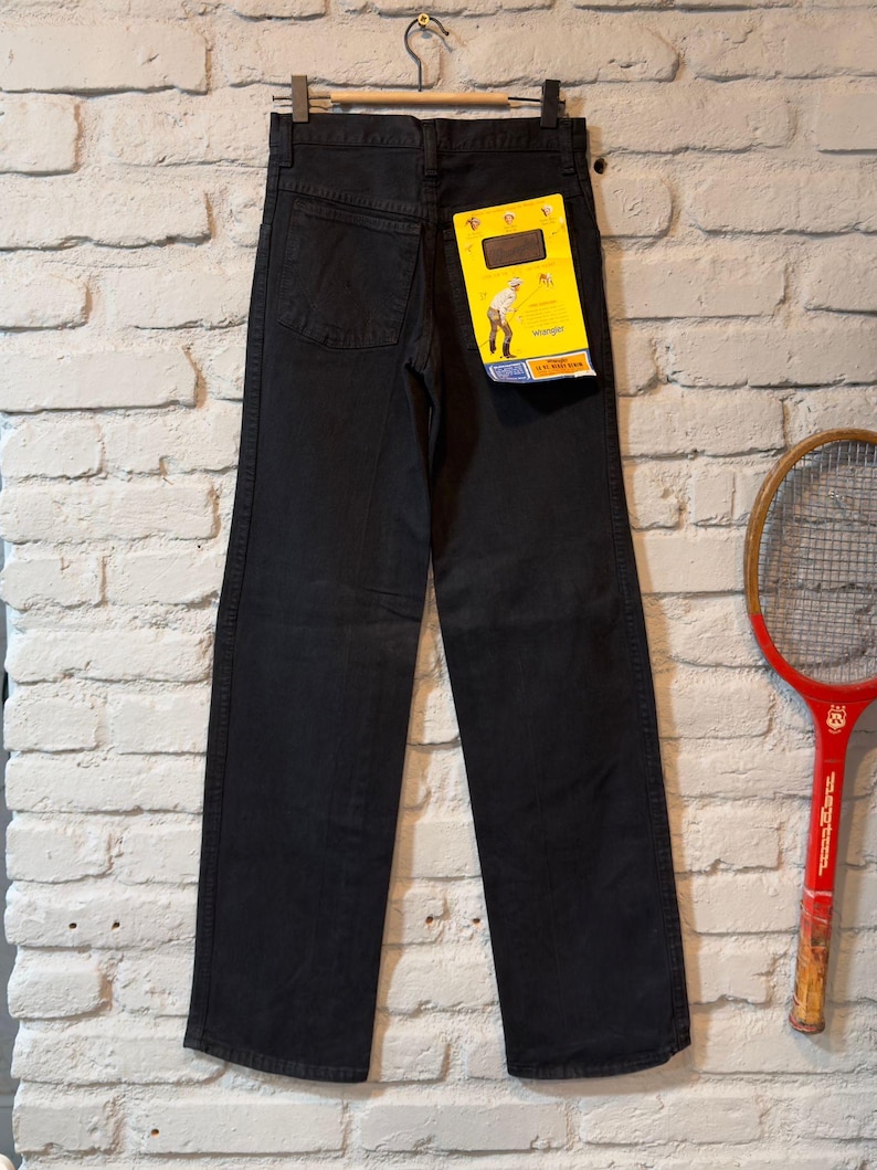 K&ouml;nnte beinhalten: Schwarze Jeans aus Denim, die an einem Metallb&uuml;gel vor einer wei&szlig;en Backsteinmauer h&auml;ngen. Die Jeans haben einen geraden Beinabschluss und ein gelbes Etikett mit einer Grafik einer Person. Ein Vintage-Tennisschl&auml;ger ist rechts zu sehen.