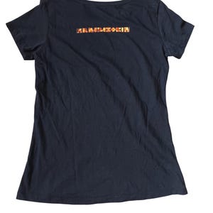 May include: Black short-sleeved t-shirt with a scoop neck. The back of the shirt features the band name "RAMMSTEIN" in a colorful, textured font. The shirt is made of a soft, dark fabric.