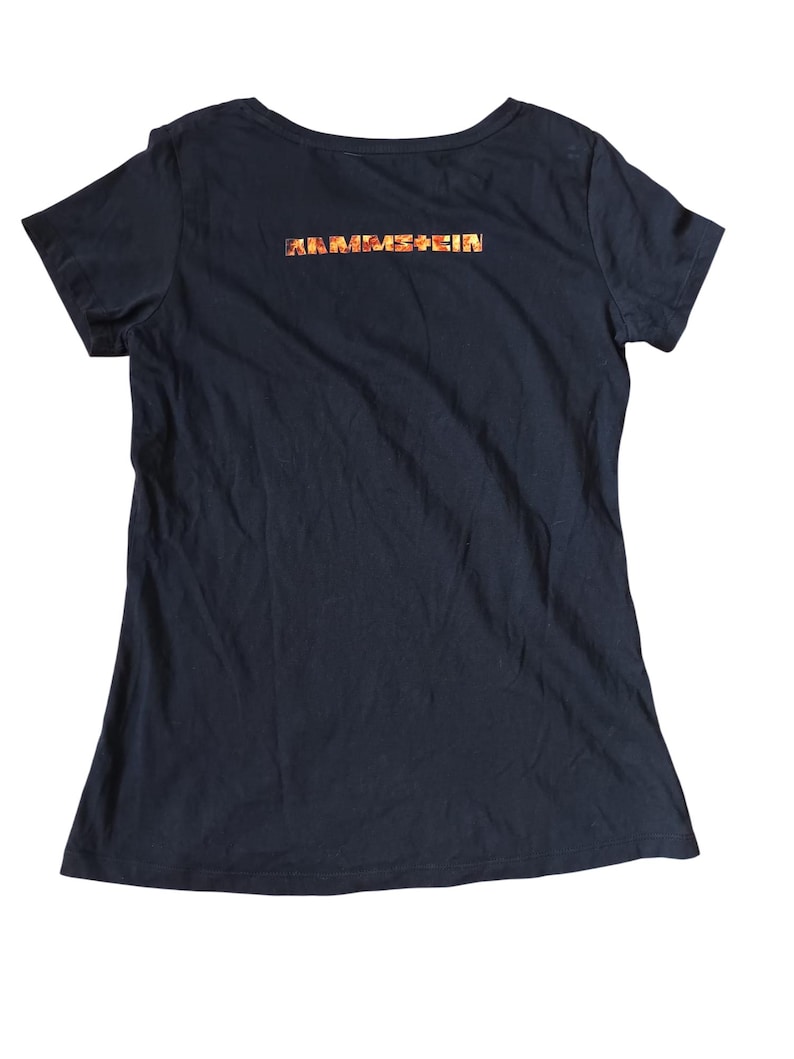 May include: Black short-sleeved t-shirt with a scoop neck. The back of the shirt features the band name "RAMMSTEIN" in a colorful, textured font. The shirt is made of a soft, dark fabric.