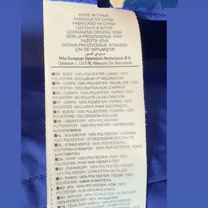 Puede incluir: Etiqueta de prenda azul con texto blanco, incluyendo "FABRICADO NA CHINA" y detalles de composici&oacute;n de la tela en varios idiomas. El logo de Nike es visible. Informaci&oacute;n sobre materiales.
