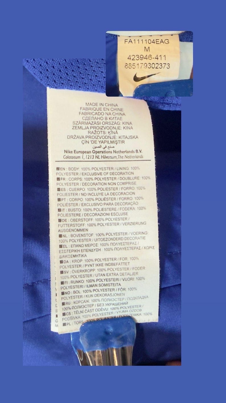 Puede incluir: Etiqueta de prenda azul con texto blanco, incluyendo "FABRICADO NA CHINA" y detalles de composici&oacute;n de la tela en varios idiomas. El logo de Nike es visible. Informaci&oacute;n sobre materiales.