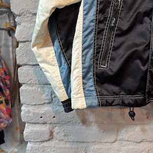Op de afbeelding: Een vintage zwarte, blauwe en cr&egrave;mekleurige jas met een ritssluiting aan de voorkant. De jas heeft een wit FILA-logo op de rits en een trekkoord aan de onderkant. De jas is gemaakt van een glanzend materiaal, mogelijk nylon.