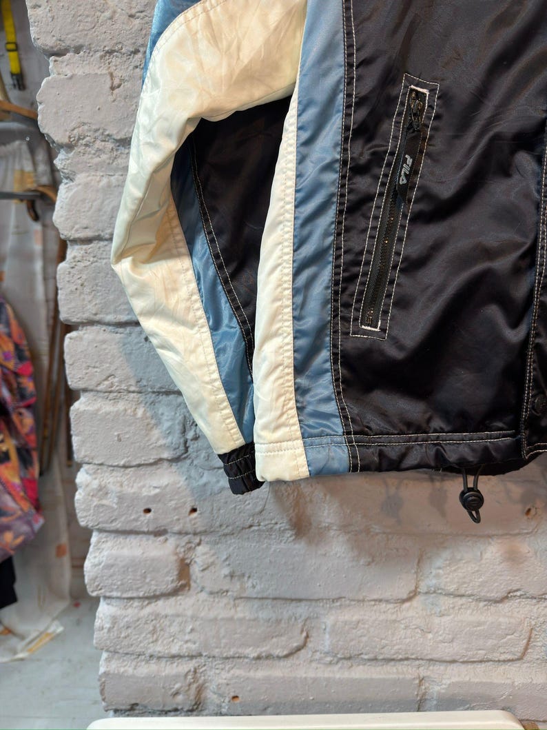 Op de afbeelding: Een vintage zwarte, blauwe en cr&egrave;mekleurige jas met een ritssluiting aan de voorkant. De jas heeft een wit FILA-logo op de rits en een trekkoord aan de onderkant. De jas is gemaakt van een glanzend materiaal, mogelijk nylon.