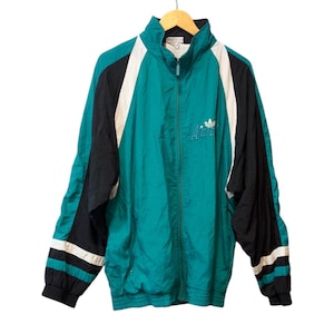 Peut inclure: Veste de survêtement vintage de couleur teal, noire et blanche, avec une fermeture éclair complète. Elle présente un col montant, des rayures blanches sur les manches et un petit logo sur la poitrine. La veste est suspendue à un cintre en bois.