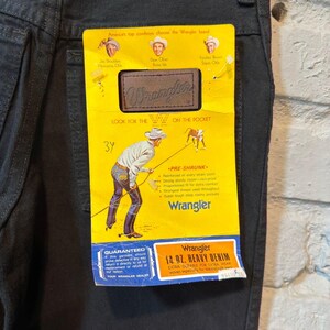 K&ouml;nnte beinhalten: Schwarze Wrangler-Jeans mit gelbem Etikett. Das Etikett zeigt das Wrangler-Logo, Cowboy-Illustrationen und den Text "14 oz. Heavy Denim". Die Jeans haben ein klassisches F&uuml;nf-Taschen-Design.