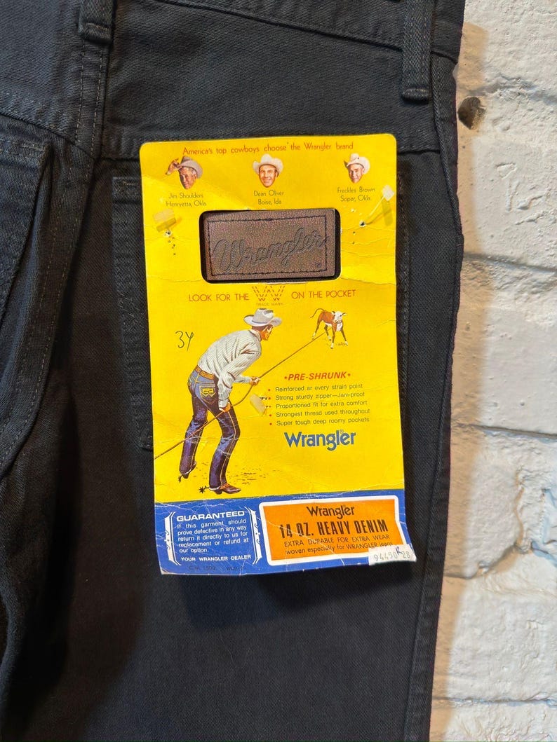 K&ouml;nnte beinhalten: Schwarze Wrangler-Jeans mit gelbem Etikett. Das Etikett zeigt das Wrangler-Logo, Cowboy-Illustrationen und den Text "14 oz. Heavy Denim". Die Jeans haben ein klassisches F&uuml;nf-Taschen-Design.