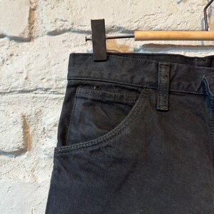 K&ouml;nnte beinhalten: Nahaufnahme einer dunkelgrauen Jeanshose, die an einem Holzb&uuml;gel h&auml;ngt. Die Hose hat ein klassisches F&uuml;nf-Taschen-Design mit einer sichtbaren Vordertasche und G&uuml;rtelschlaufen. Der Stoff scheint ein robustes, strapazierf&auml;higes Material zu sein.